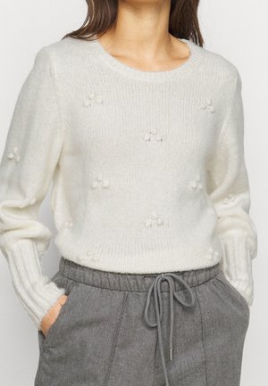 Kobieta ubrana w biały sweter z dzianiny z małymi kuleczkami pomponów oraz szare spodnie z sznurkiem w talii, trzymająca ręce w kieszeniach.