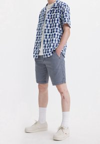 Chemise à manches courtes à carreaux bleu et blanc, associée à un short gris. Des chaussettes blanches et des chaussures beiges complètent la tenue.
