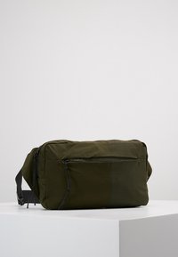 Olivově zelená textilní taška přes rameno s přední kapsou na zip, nastavitelným popruhem a hladkým povrchem. Obdélníkový tvar s minimalistickým designem.