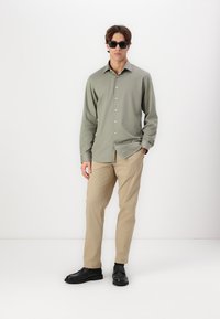 Lys grønn skjorte med knapper og rett krage, beige bukser og svarte sko. Modell med mørke solbriller, som står mot en hvit bakgrunn.