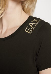 Svart åtsittande t-shirt med rund halsringning, med ett guld "EA7 Emporio Armani"-logga på axeln. Mjuk, slät tyg med subtil textur.