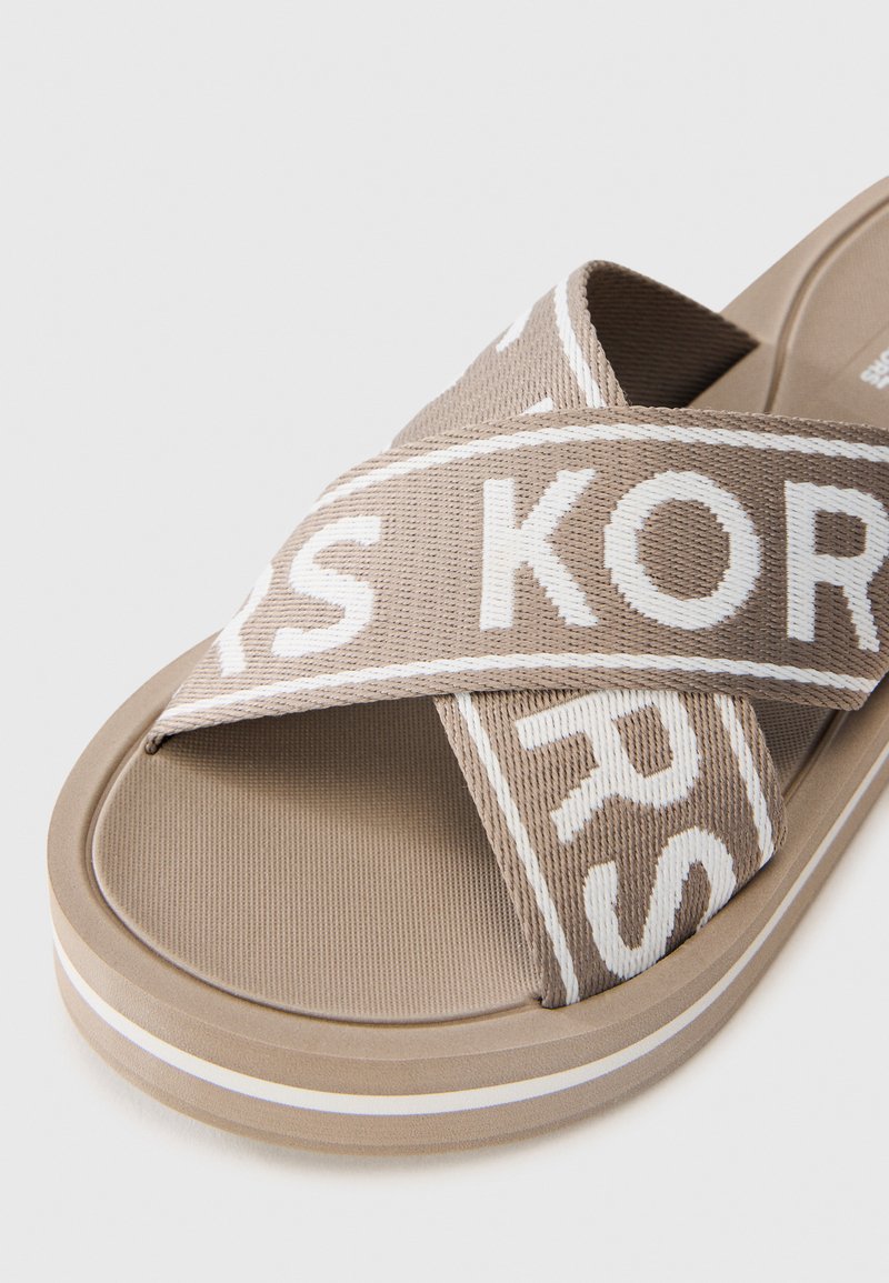 Chanclas marrones con tiras entrelazadas que presentan texto en blanco con el logo. Tiras de tela texturizada y una suela plana y suave con una franja blanca de acento.