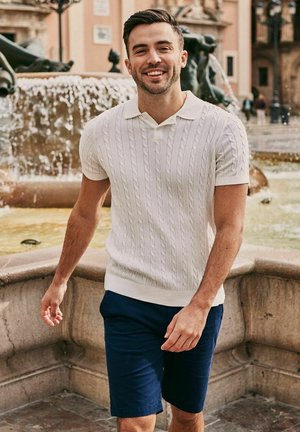 Lachende man in een wit kortärmelig vest en marineblauwe korte broek wandelt vlak bij een stenen fontein op een historisch stadsplein.