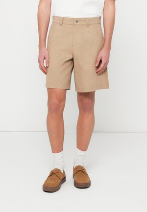 Χακί σορτς με επίπεδη εμπρός όψη, πλαϊνές τσέπες και κουμπωτό κλείσιμο. Φορεμένα με καφέ σουέντ παπούτσια και λευκές κάλτσες.