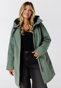 Groene waterdichte parka met een capuchon, voorzien van een zwarte voering, voorknopsluitingen en zijzakken met rits. Gecombineerd met zwarte jeans.