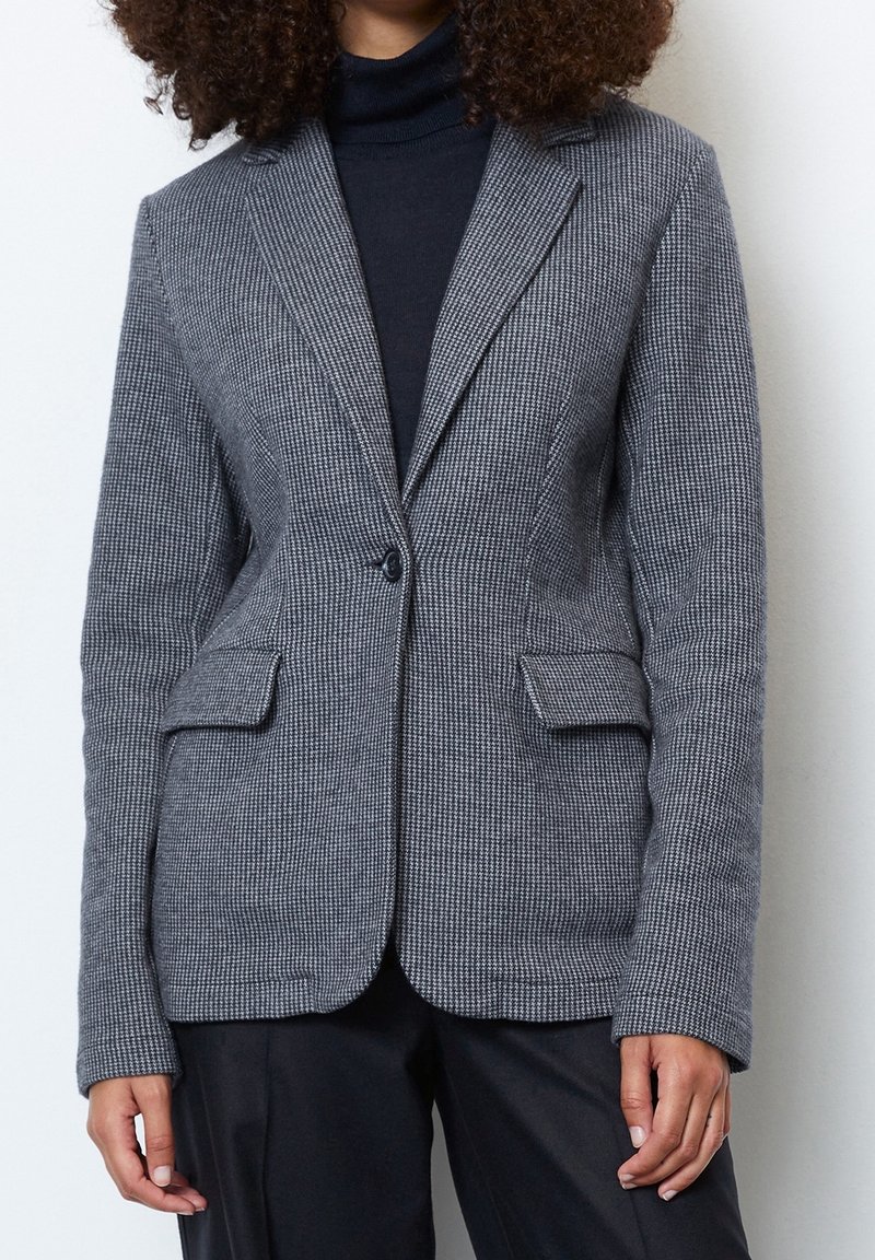 Femme portant un blazer gris à motif pied-de-poule ajusté sur un col roulé noir et un pantalon noir, debout devant un fond blanc uni.