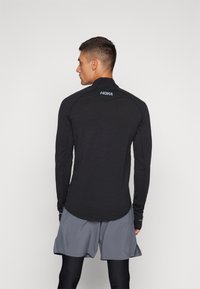 Haut de sport noir à manches longues avec un col haut, arborant un logo discret au dos. Associé à un short gris pour un look sportif.