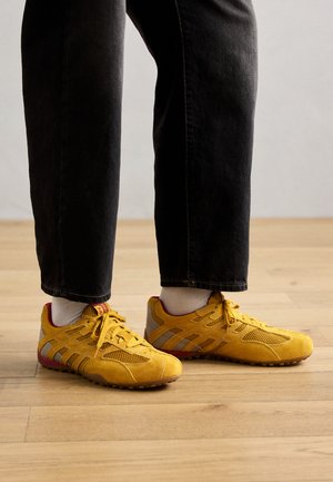 Scarpe da ginnastica gialle in materiali mesh e suede. Presentano accenti grigi, suole piatte e un design leggero. Indossate con pantaloni neri.