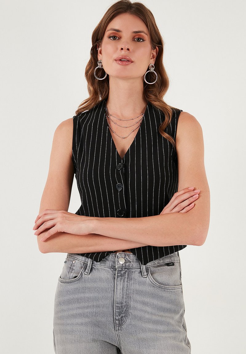 Čierna bezrukávová blúzka s vertikálnymi bielymi pruhmi, s výstrihom do V, gombíkovým zapínaním a textúrovanou tkaninou; skombinovaná s svetlošedými džínami s vysokým pásom.