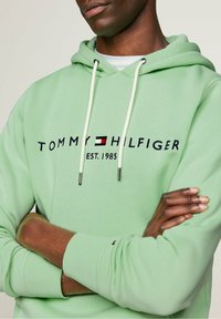 Felpa verde chiaro con cappuccio e lacci, con la scritta "TOMMY HILFIGER EST. 1985" e logo. Texture morbida, design minimalista, polsini a costine.