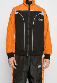 Black and orange athletic jacket with white accents, zippered front, and high collar. Smooth texture and elastic cuffs. Logo on chest.