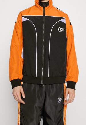 Chaqueta deportiva negra y naranja con acentos blancos, cierre en la parte frontal y cuello alto. Textura suave y puños elásticos. Logo en el pecho.