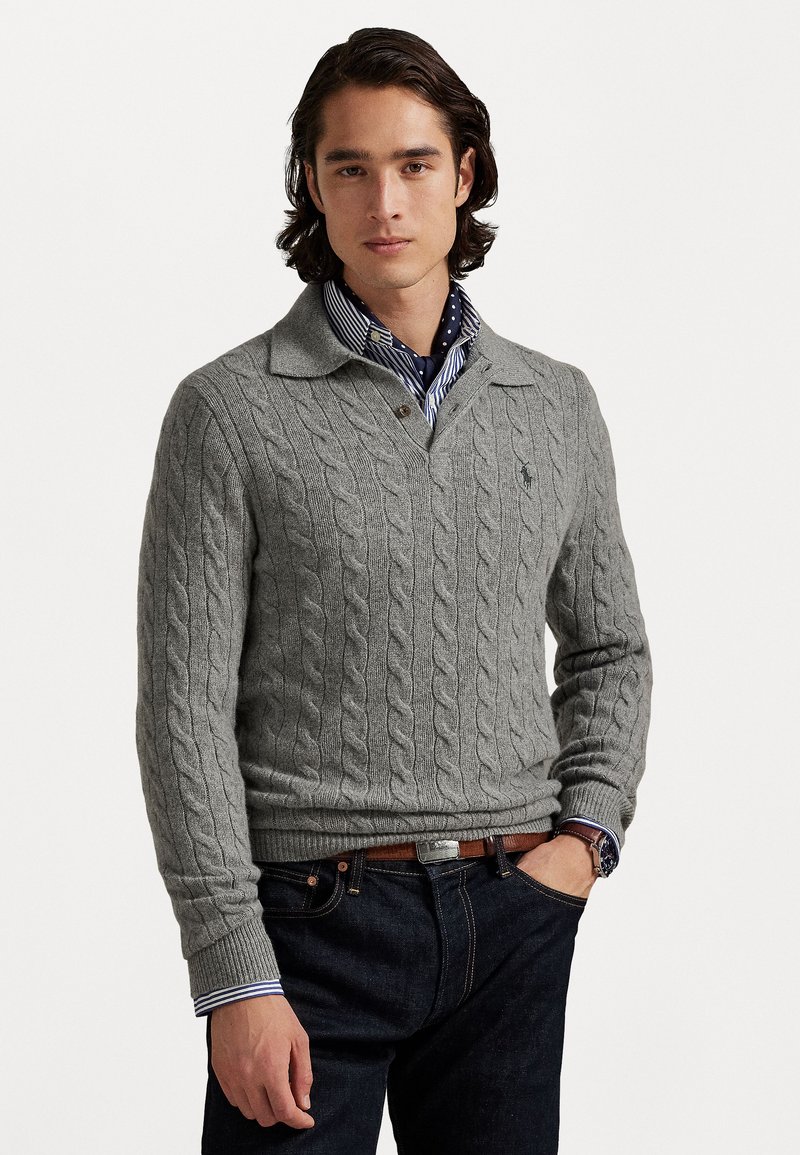Pull en maille grise à col, avec un petit logo et des poignets côtelés, porté avec une chemise rayée bleu marine et blanche en dessous.