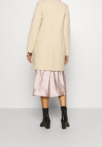 Cappotto beige con taglio dritto, in tessuto misto lana, abbinato a una gonna in satin floreale rosa e stivali neri alla caviglia con tacco blocco.