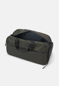 Mala de viagem verde-oliva com uma superfície suave e texturizada, alças de tecido, fecho superior com zíper e uma marca ao longo do lado. O interior é azul escuro.