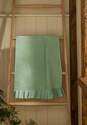 Serviette vert clair avec bord froncé suspendue à une échelle en bois contre un mur beige avec une poutre en bois apparente.