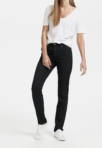 Mujer vistiendo pantalones ajustados negros, camiseta blanca de cuello en V de manga corta y zapatillas blancas, de pie contra un fondo blanco liso.