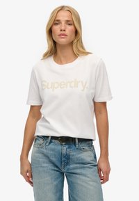 Hvid kortærmet t-shirt med præget "Superdry." logo i beige, båret med lyseblå denimjeans og et sort bælte.