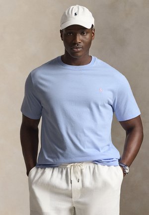 Muž oblečený v svetlomodrej polokošeli, bielych nohaviciach so šnúrkou, bielej čiapke a hodinkách, stojac pred jednoduchým béžovým pozadím.