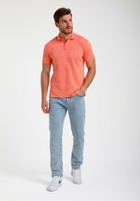 Koralna polo majica z teksturirano površino, z zapenjanjem na tri gumbe, v kombinaciji z svetlo modrimi kavbojkami in belimi supergami, ki vključujejo subtilno podrobnost logotipa.