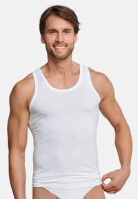 Witte geribde tanktop van katoen, met brede bandjes en een aansluitend ontwerp. Het model glimlacht en toont de pasvorm van de top.