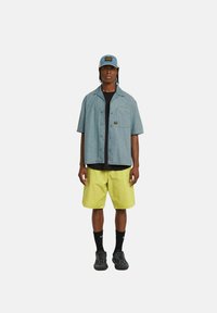 Camisa de botón de manga corta azul claro, camiseta negra, pantalones cortos amarillos, calcetas negras y zapatillas, con una gorra que lleva un parche con un logo.