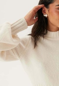 Pull crème avec des poignets et un col côtelés ; texture douce et coupe décontractée ; porté avec une boucle d'oreille en forme de cœur ; main partiellement visible.