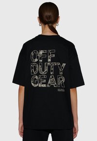 Person med hästsvans som bär en svart oversize-T-shirt med texten "OFF DUTY GEAR" i stora mönstrade bokstäver på baksidan.