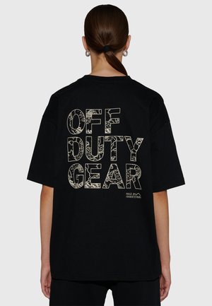 Person med hästsvans som bär en svart oversize-T-shirt med texten "OFF DUTY GEAR" i stora mönstrade bokstäver på baksidan.