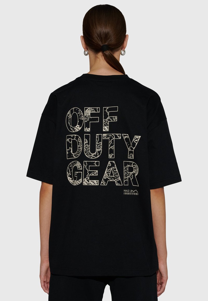 Person med hästsvans som bär en svart oversize-T-shirt med texten "OFF DUTY GEAR" i stora mönstrade bokstäver på baksidan.