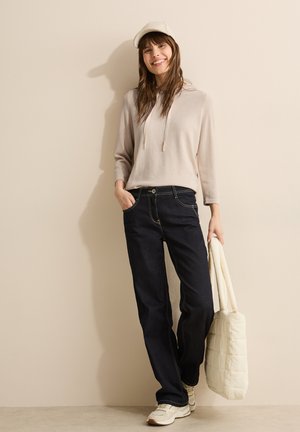 Felpa beige con lacci, pantaloni a gamba larga in denim scuro, sneaker chiare e borsa a tracolla trapuntata color crema, su uno sfondo neutro.