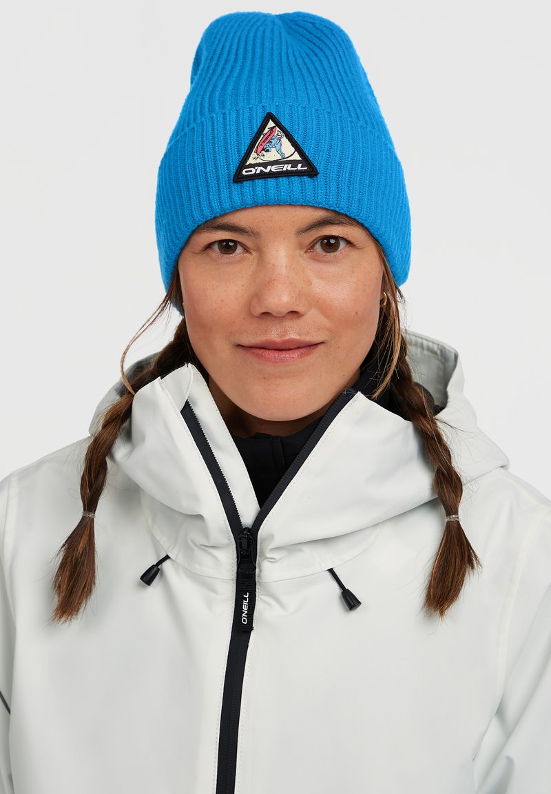 Blå ribbet strikket beanie med et trekantet logomærke, parret med en hvid jakke med lynlås og hætte, der har sorte detaljer.