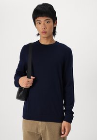 Tamsiai mėlynas, ilgam rankovėms megztinis su apvaliu iškirpimu, pagamintas iš minkštos medžiagos. Derinamas su šviesiai rudos spalvos korduroy kelnėmis ir mažyte juoda rankine.