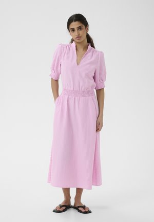 Frau trägt ein hellrosa gestreiftes Midikleid mit Puffärmeln, geraffter Taille, Taschen und schwarzen flachen Sandalen, steht vor einem weißen Hintergrund.