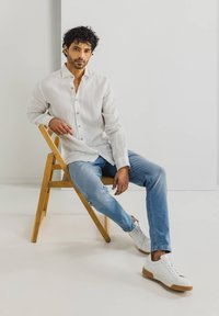 Weißes Leinenhemd mit Knopfleiste, hellblaue Jeans und weiße Sneakers mit Gummisohlen; sitzt auf einem Holzstuhl vor einem neutralen Hintergrund.