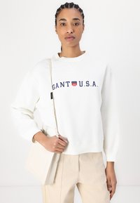 Sweatshirt en polaire blanche avec "GANT U.S.A." brodé en marine et un design graphique. Le modèle tient un sac bandoulière beige clair.