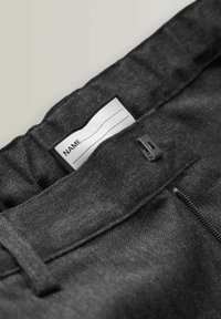 Gros plan de la ceinture d'un pantalon gris foncé montrant une bande élastique, des passants pour ceinture, une pince noire et une étiquette de nom blanche cousue avec des lignes pointillées.