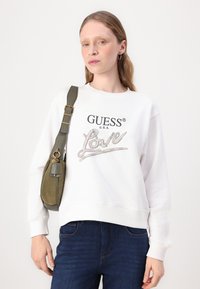 Felpa bianca con la scritta "GUESS U.S.A." e "Love" in lettere scintillanti. Abbinata a una borsa a tracolla verde oliva. Jeans scuri.