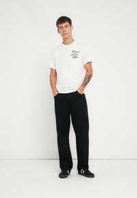 Tricou alb din bumbac cu imprimeu grafic negru, combinat cu pantaloni negri largi și adidași negri cu dungi albe. Fundal uniform.