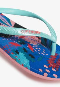 Flip-flop med en turkis rem, der har en blomsterblå og pink mønstret fodseng og en lys pink sål. "SUPERDRY" logoet i rød.