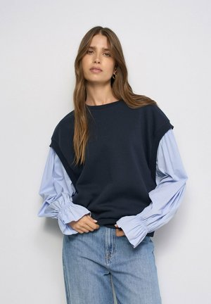 Mujer con cabello largo y castaño que lleva una blusa sin mangas negra sobre una blusa azul claro con mangas abullonadas y vaqueros azul claro, de pie contra un fondo liso.