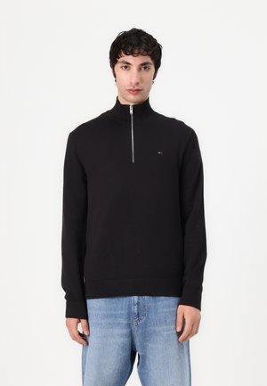 Zwarte gebreide pullover met een hoge kraag en een halve rits. Kenmerkt zich door een klein logo op de borst en ribgebreide manchetten en zoom.
