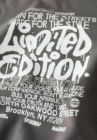Donkere stof met witte tekst in graffiti-stijl met de woorden "Limited Edition," straatmode slogans, adres in Brooklyn, NY, en een klein wereldbol icoon.
