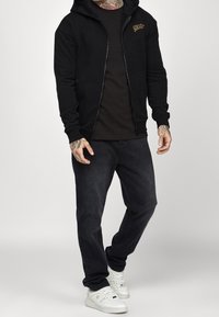Sudadera negra con cremallera frontal, tejido texturizado, dos grandes bolsillos, usada sobre una camiseta negra y combinada con jeans oscuros y zapatillas blancas.