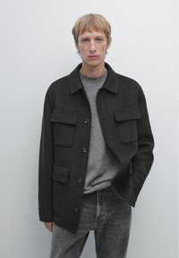 Donkergrijze wollen jas met een kraag, twee borstzakken en knoopsluiting, gedragen over een lichtgrijze trui en donkere spijkerbroek.