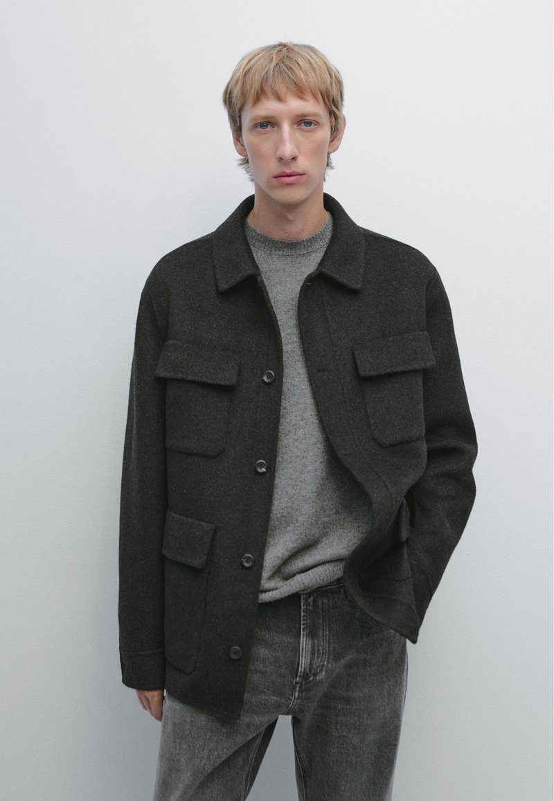 Donkergrijze wollen jas met een kraag, twee borstzakken en knoopsluiting, gedragen over een lichtgrijze trui en donkere spijkerbroek.