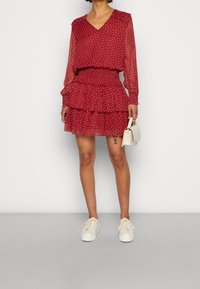 Robe mini rouge à volants avec un décolleté en V, des manches longues transparentes et un imprimé floral. Associée à des baskets blanches et un petit sac à main.