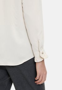 Crèmekleurige blouse met lange mouwen, voorzien van knoopmanchetten en geruimte randen. Gekleed met donkergrijze broek. Soepele textuur.