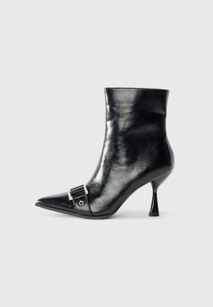 Black ankle boot made of glossy leather with a pointed toe, featuring a silver buckle detail and a unique triangular heel design.