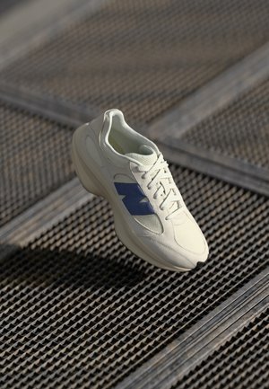 Baskets blanches avec logo latéral bleu flottant au-dessus d'une surface en grille métallique sous une lumière naturelle douce.